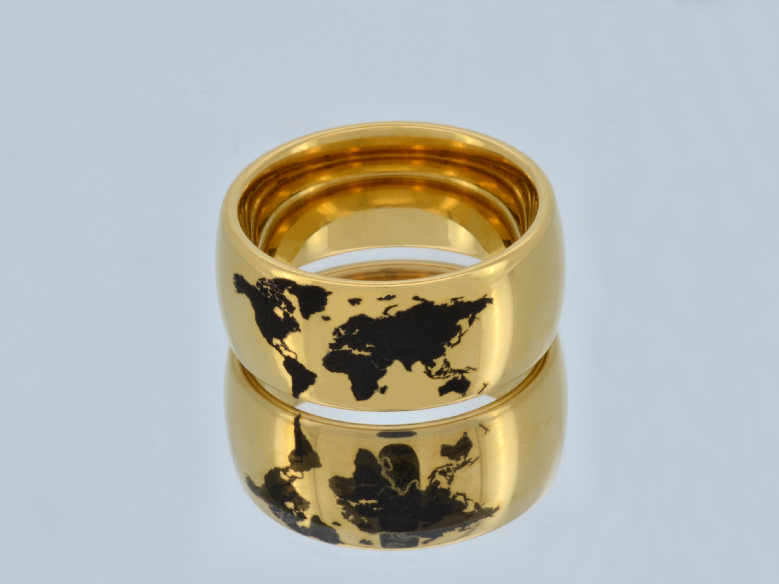 Gold tungsten ring with a black engraved world map, displayed on a reflective surface showing a mirrored view of the design