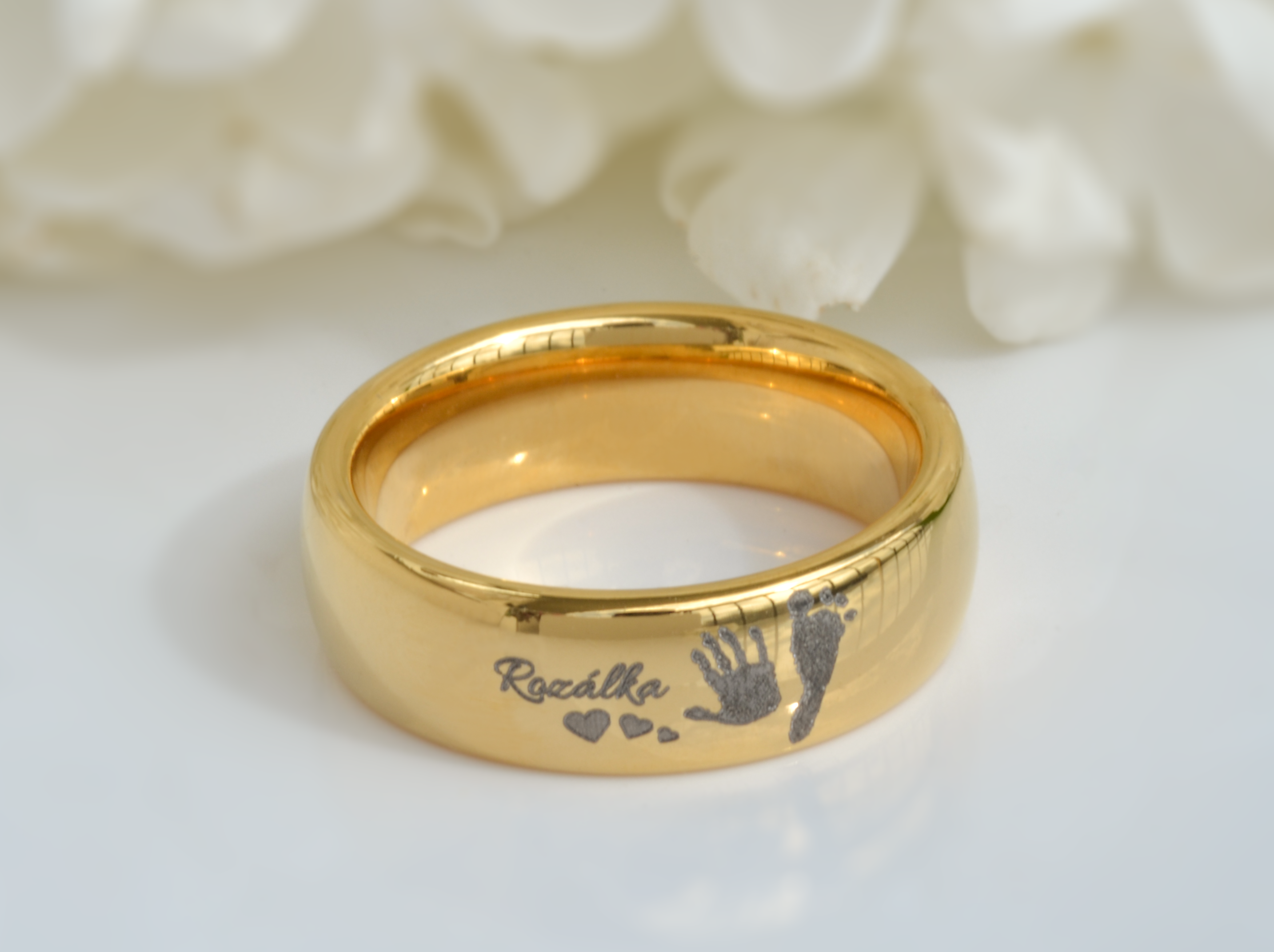 A golden tungsten ring named 'Eternal Imprint' displayed against a soft, white floral background. The ring features a personalized engraving of a child's handprint and footprint alongside small hearts, capturing a sentimental and enduring memory in a durable piece of jewelry.