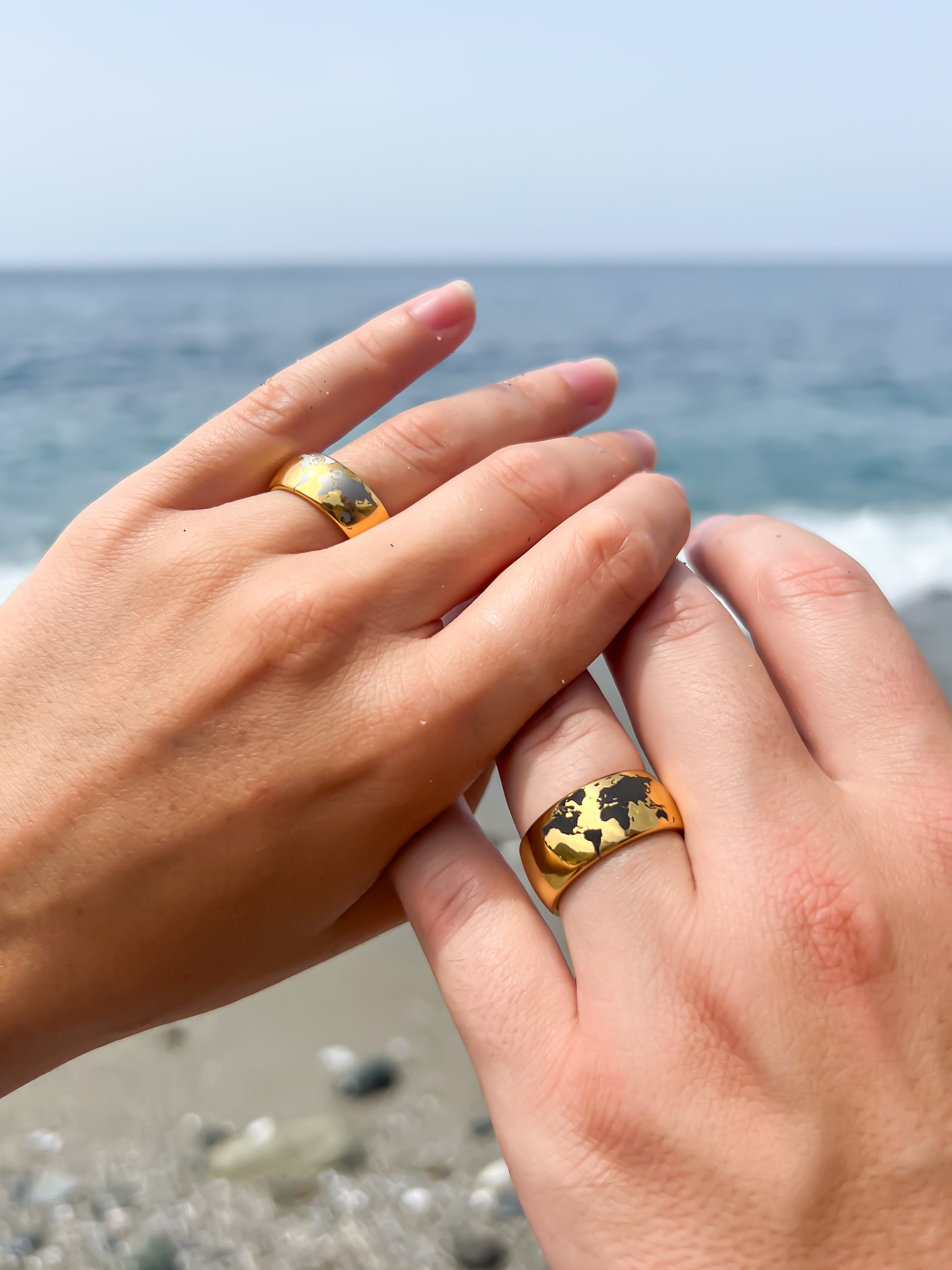 Two hands wearing matching gold tungsten rings with black world map design, photographed by the sea on a sandy beach.