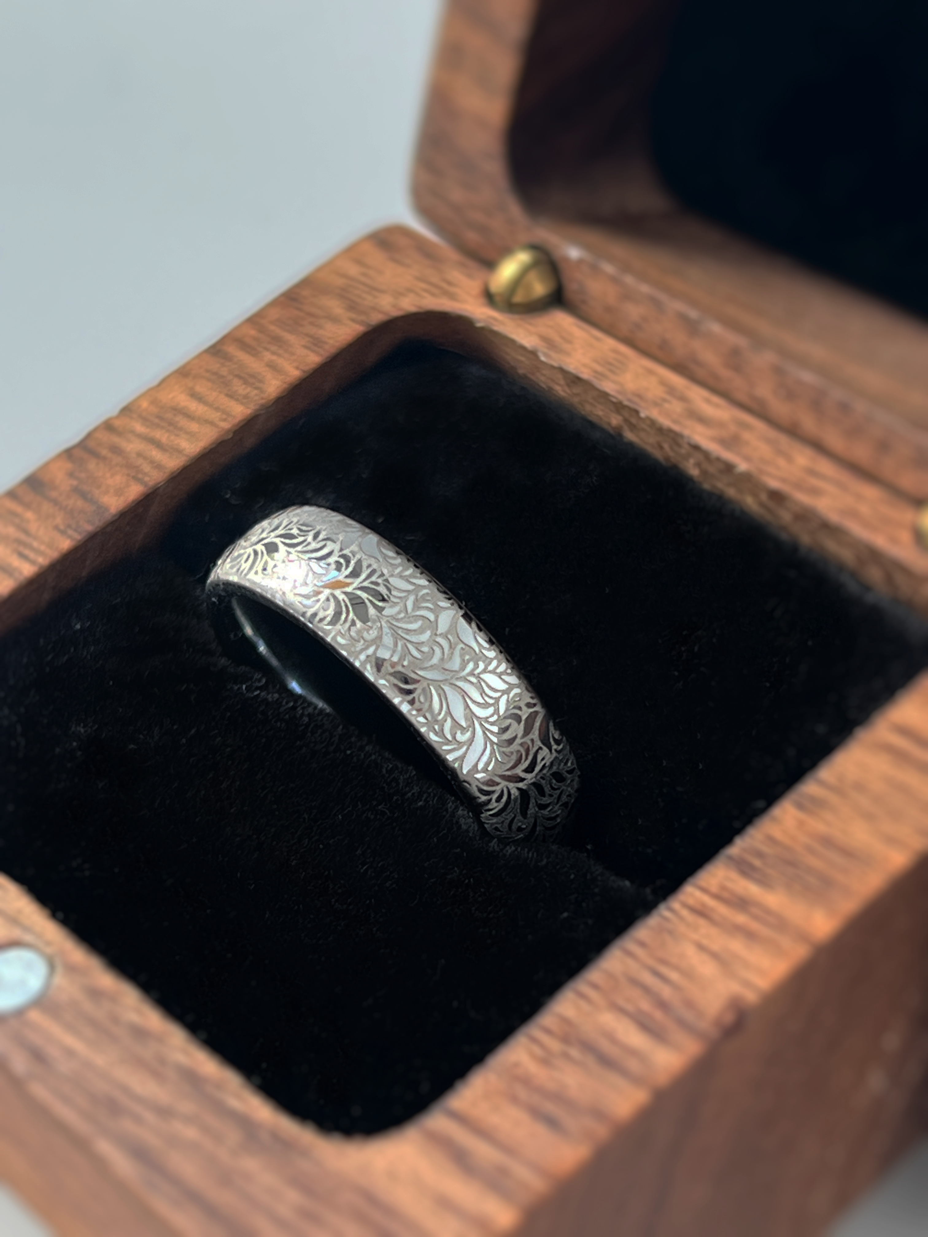 Elegant white gold tungsten displayed inside wooden box. The ring has intricate silver floral engraving, featuring a polished shiny finish and delicate patterns. Durable and stylish, this engraved tungsten wedding or engagement band for her or him