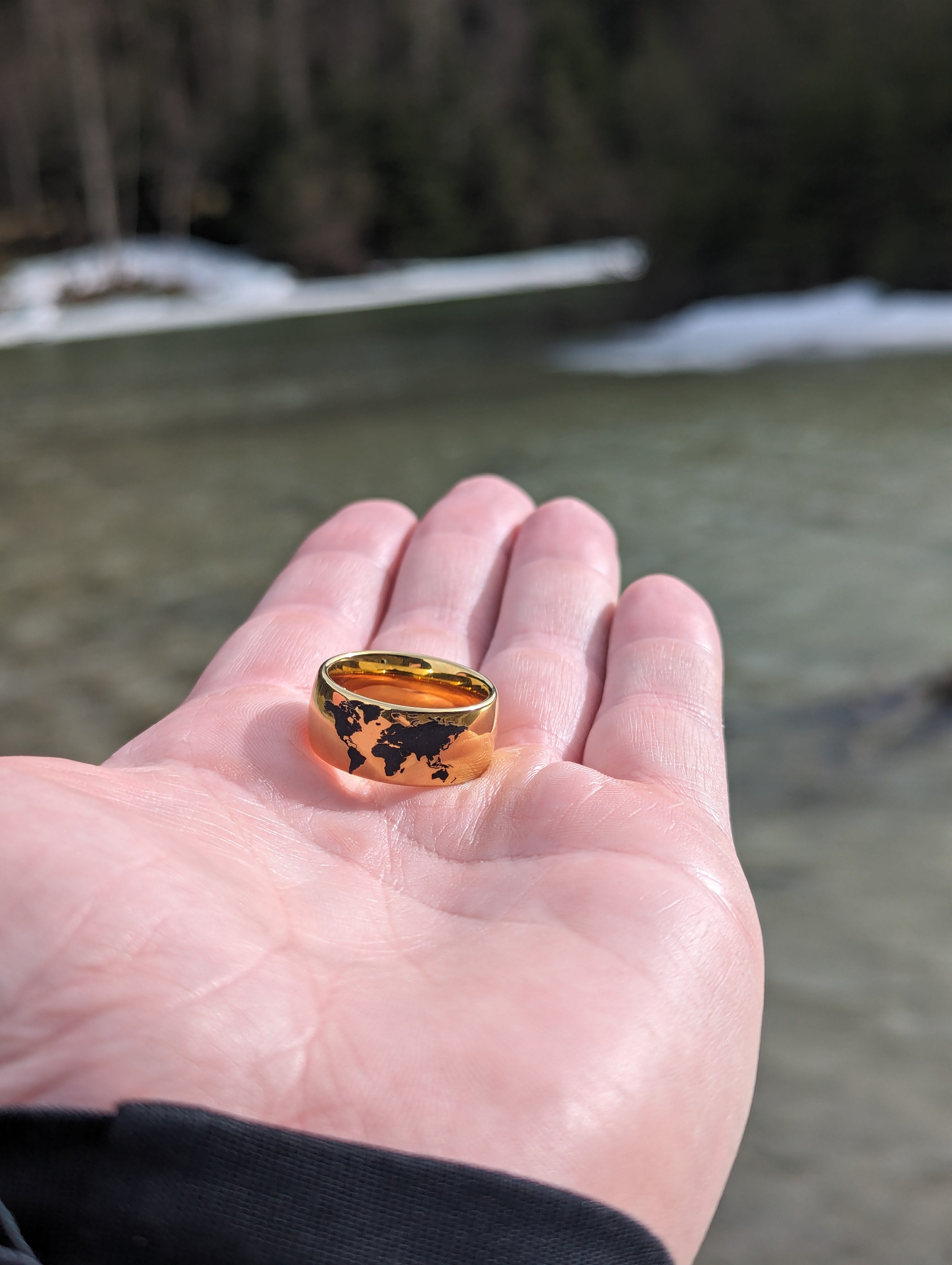 Hand holding a gold tungsten ring with a black world map design, photographed outdoors by a river with a blurred natural background.