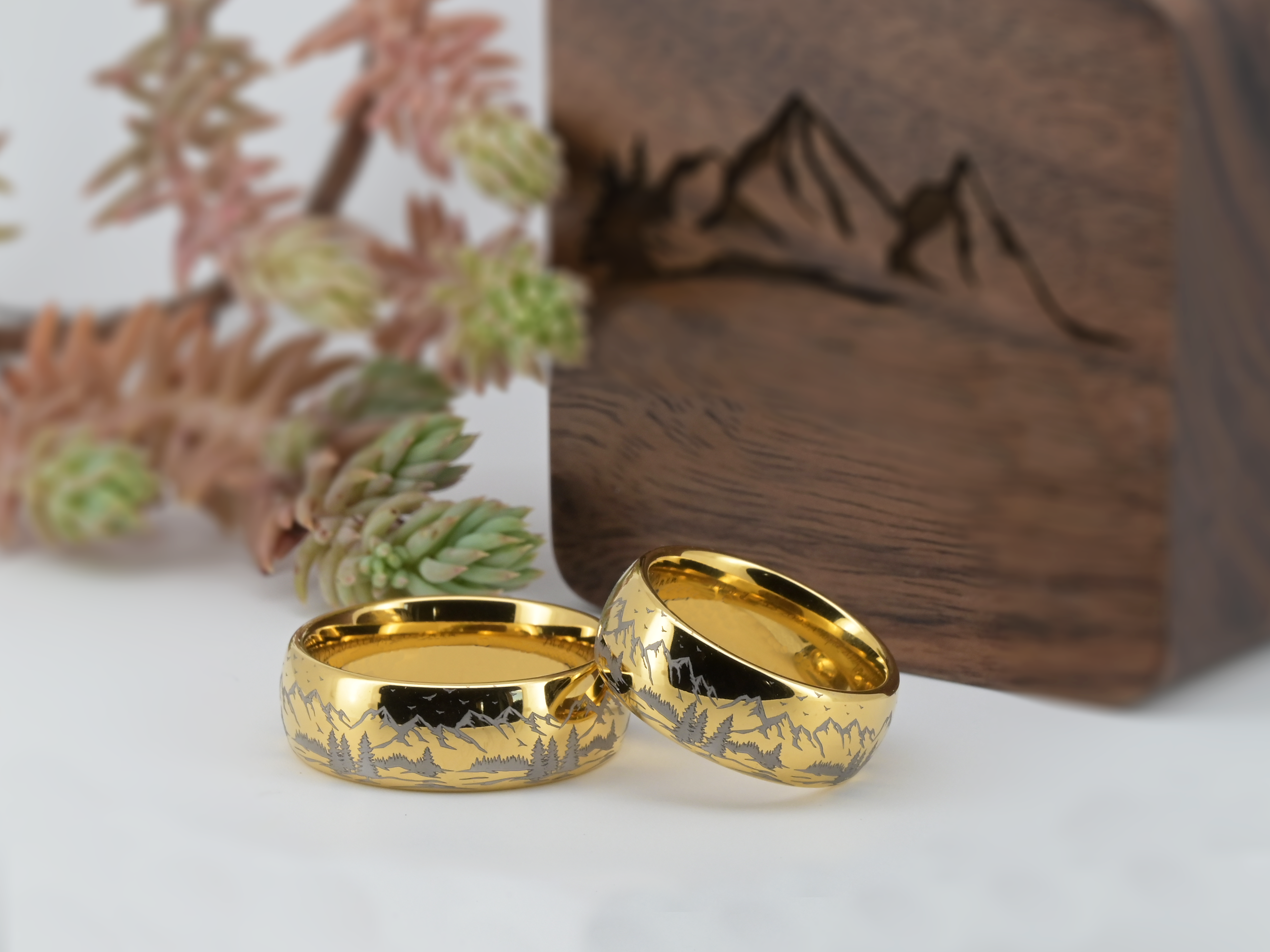 Two gold tungsten rings with mountain engraving displayed next to a wooden ring box and natural decor
