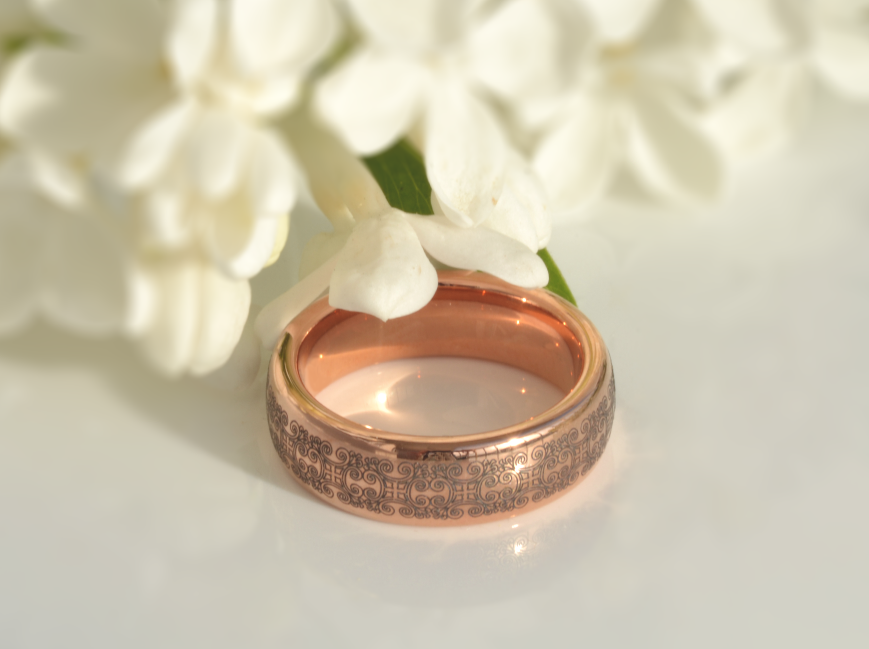 Rose gold tungsten ring with intricate engravings and rose gold interior, resting beside white flowers on a soft, glowing background.