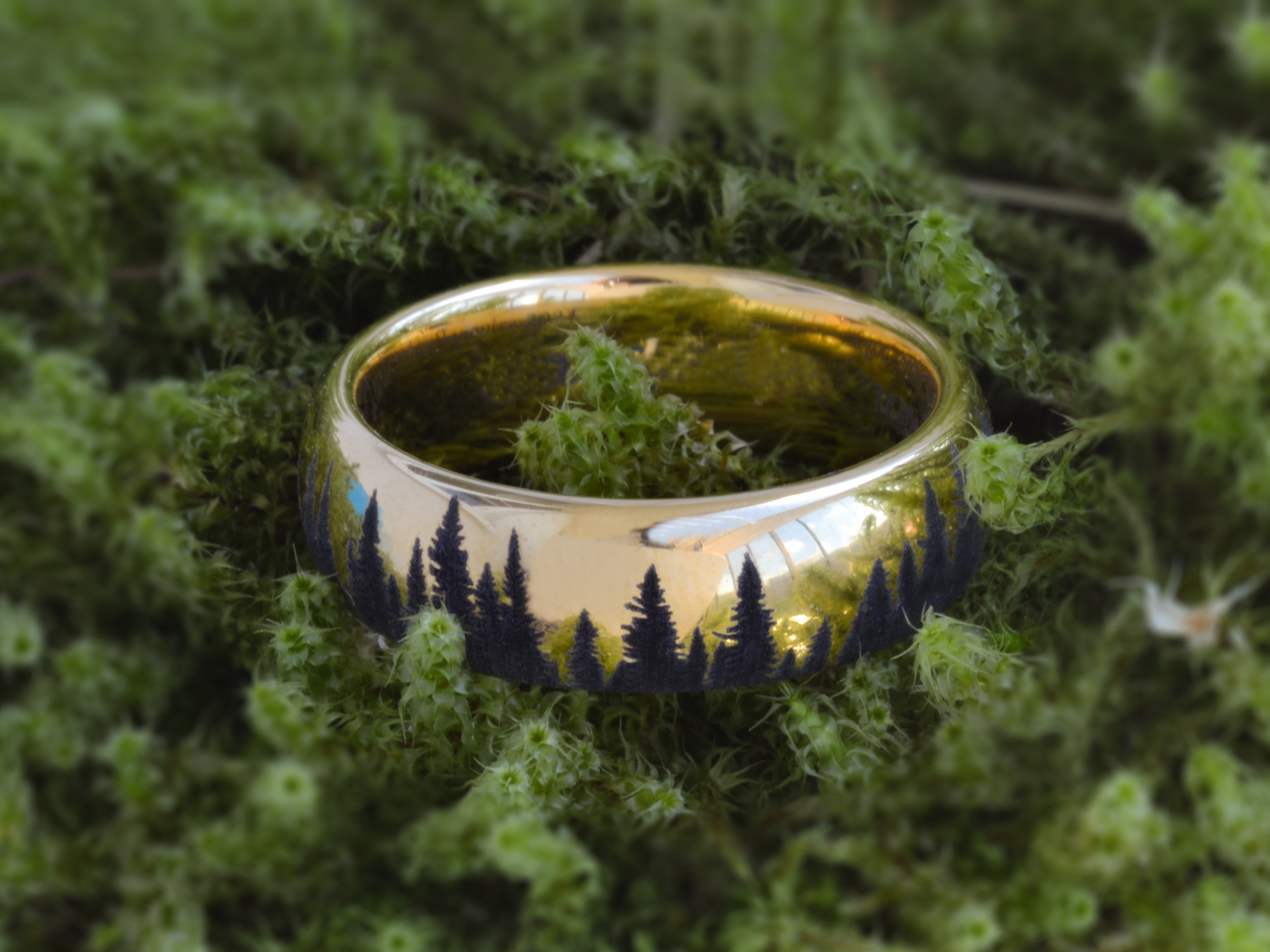 Gold tungsten ring with an engraved silhouette of an evergreen forest, placed on a piece of bark with moss, showcasing the nature-inspired design