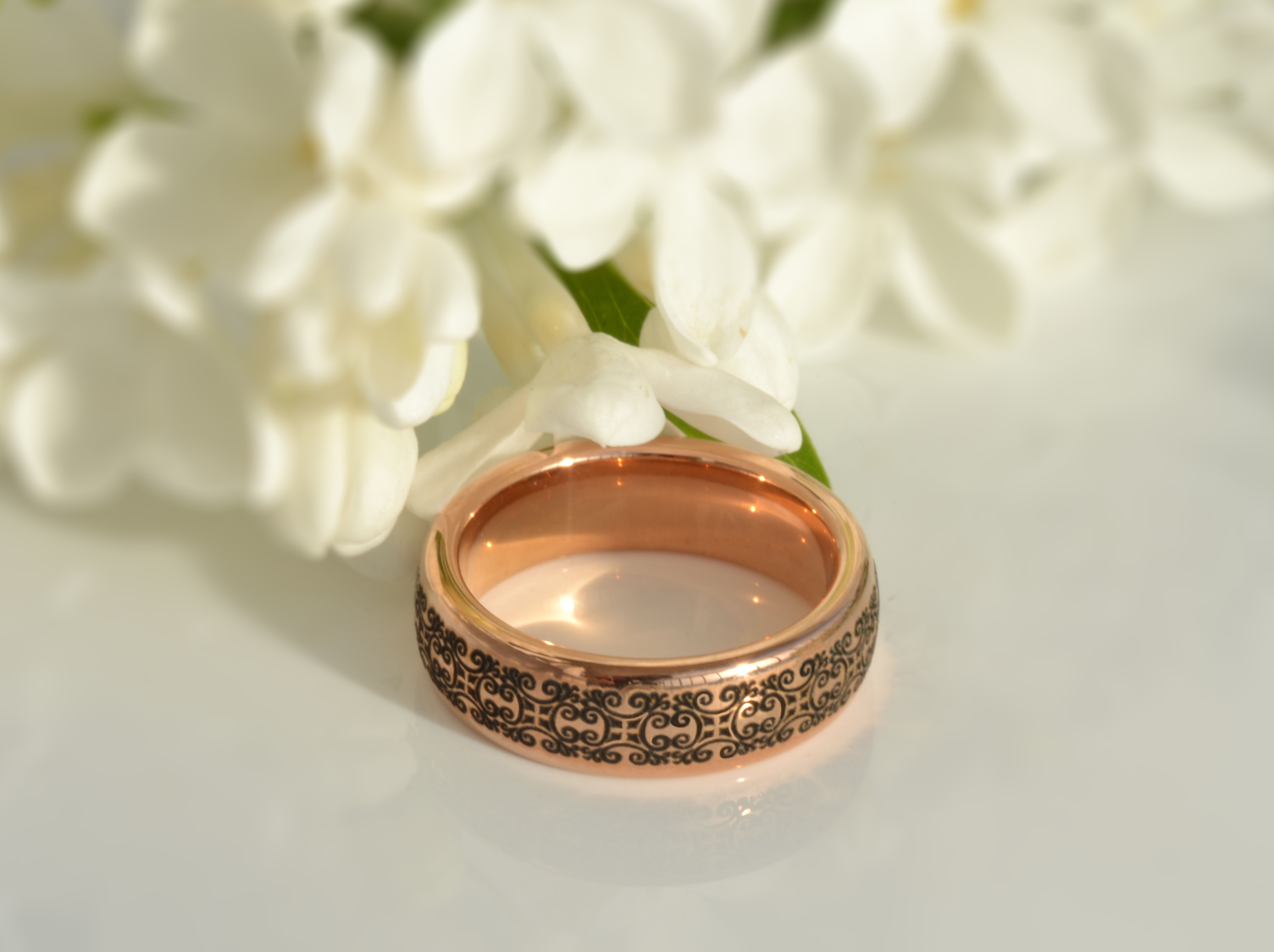 Close-up of a rose gold tungsten ring with an antique black swirl pattern, placed in front of white flowers.