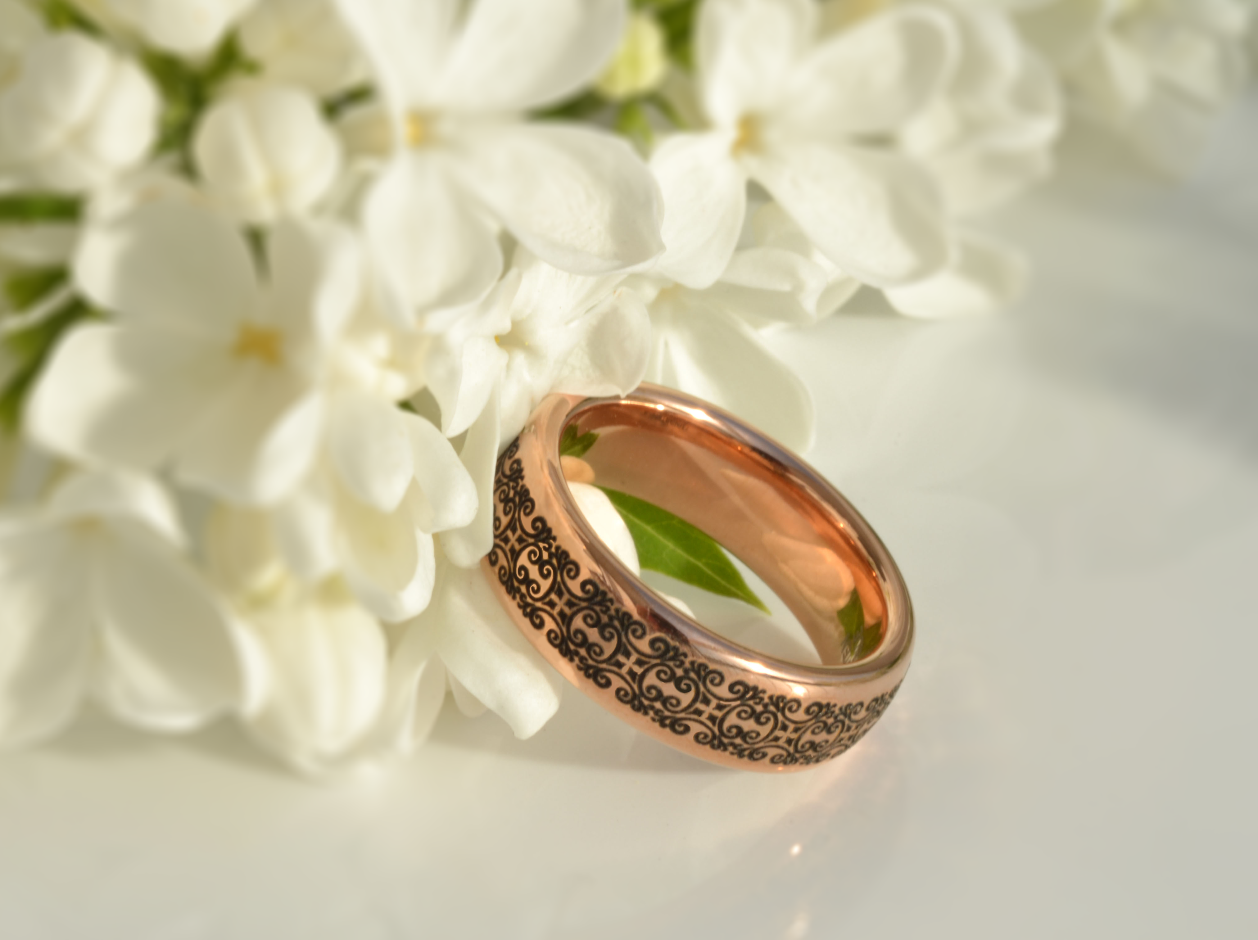 Rose gold tungsten ring with an intricate black swirl design, surrounded by white flowers.