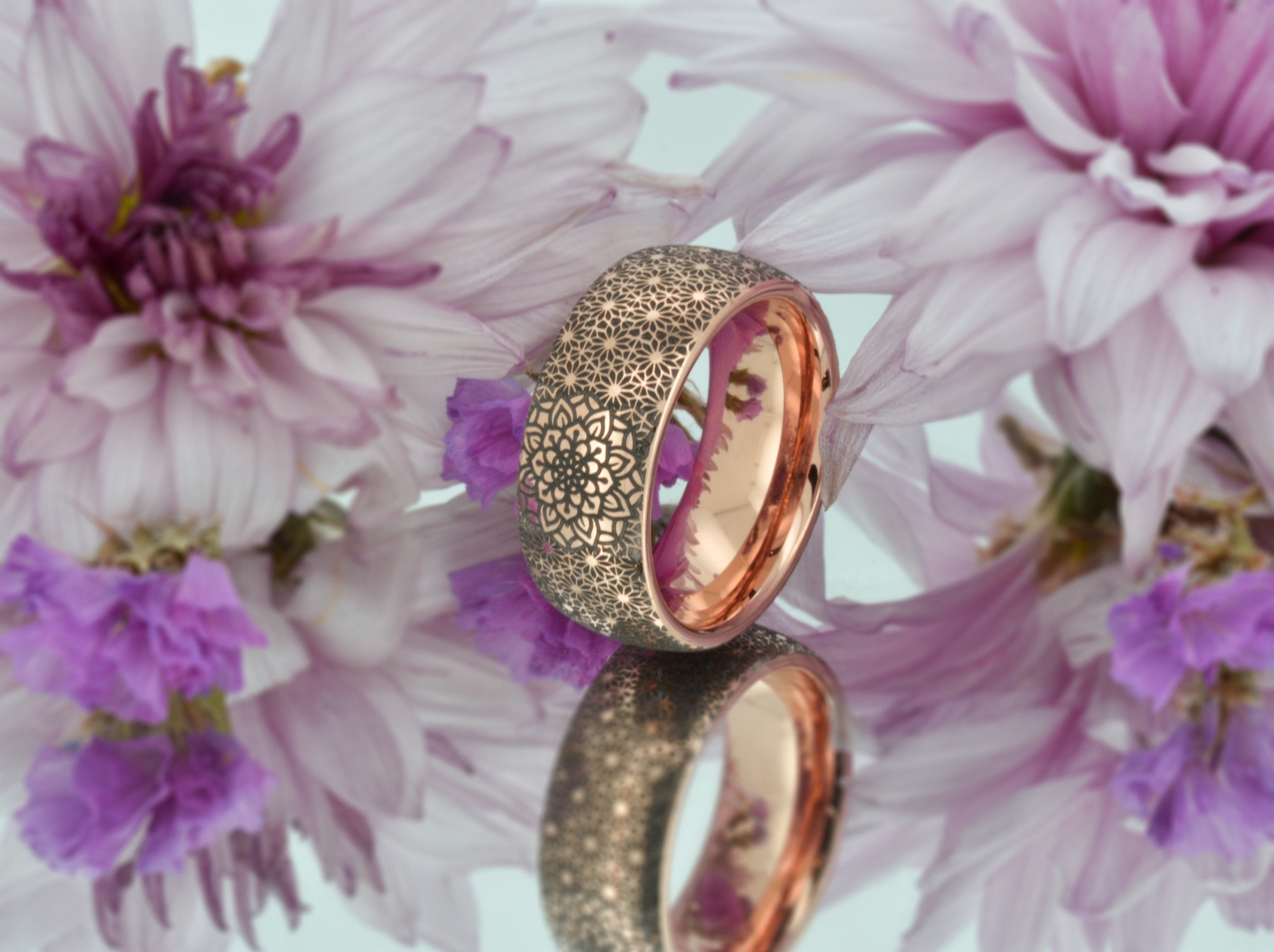 Rose gold tungsten ring with boho floral mandala design and a shiny rose gold interior, artistically displayed among soft purple flowers, reflecting on a mirrored surface.