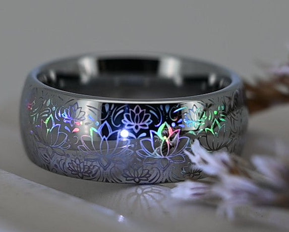 A silver-toned band ring with a textured, hammered finish and small colorful inlay details, resting on a white display block next to dried brown plant stems.