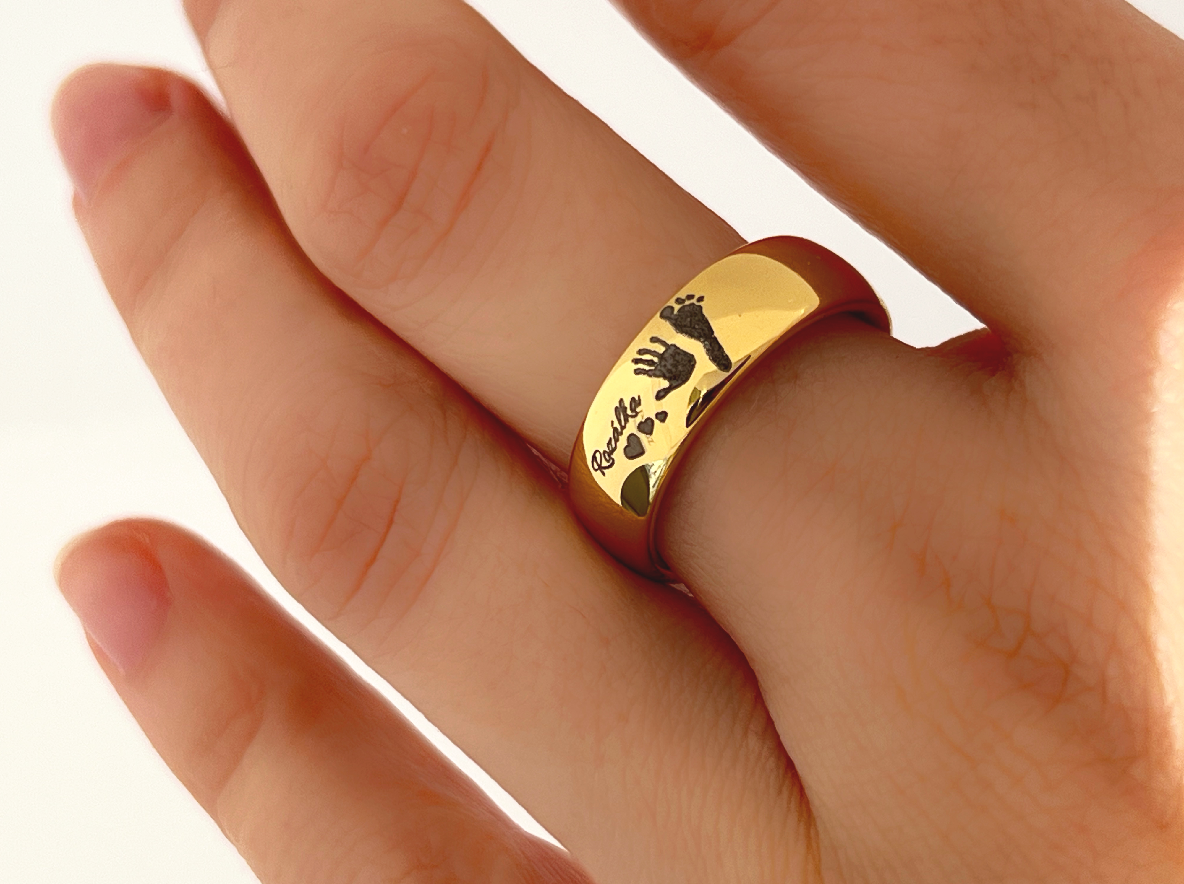 Eine Hand trägt einen goldenen Wolframring namens Eternal Imprint, in den ein echter silbergrauer Hand- und Fußabdruck eines Kindes eingraviert ist. Die personalisierten Gravuren auf dem Ring machen ihn zu einem bedeutungsvollen Schmuckstück, das die dauerhafte Verbindung zwischen Eltern und Kind symbolisiert.