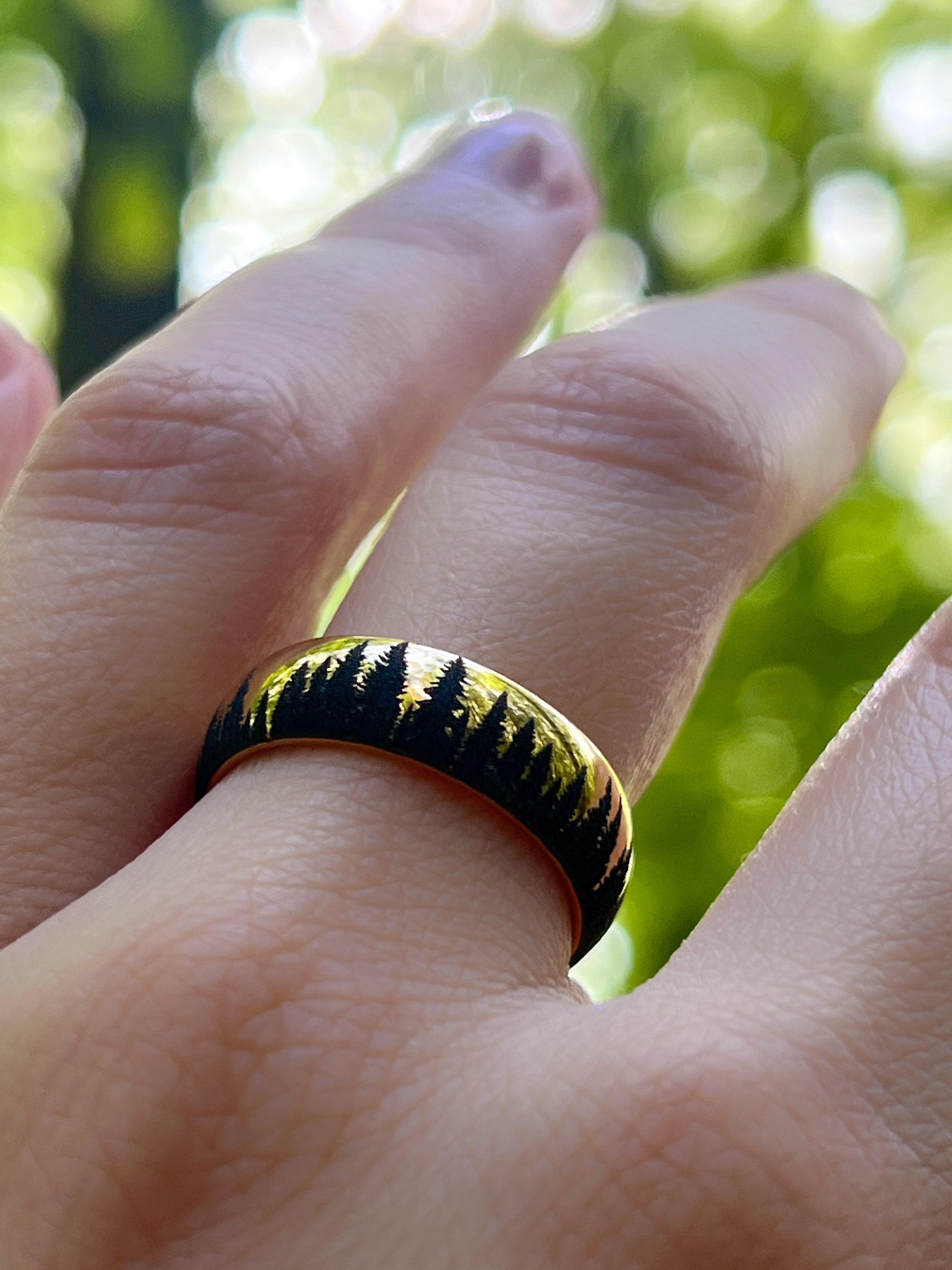 Gold tungsten ring with an engraved silhouette of an evergreen forest, worn on a finger, showcasing the detailed forest design.