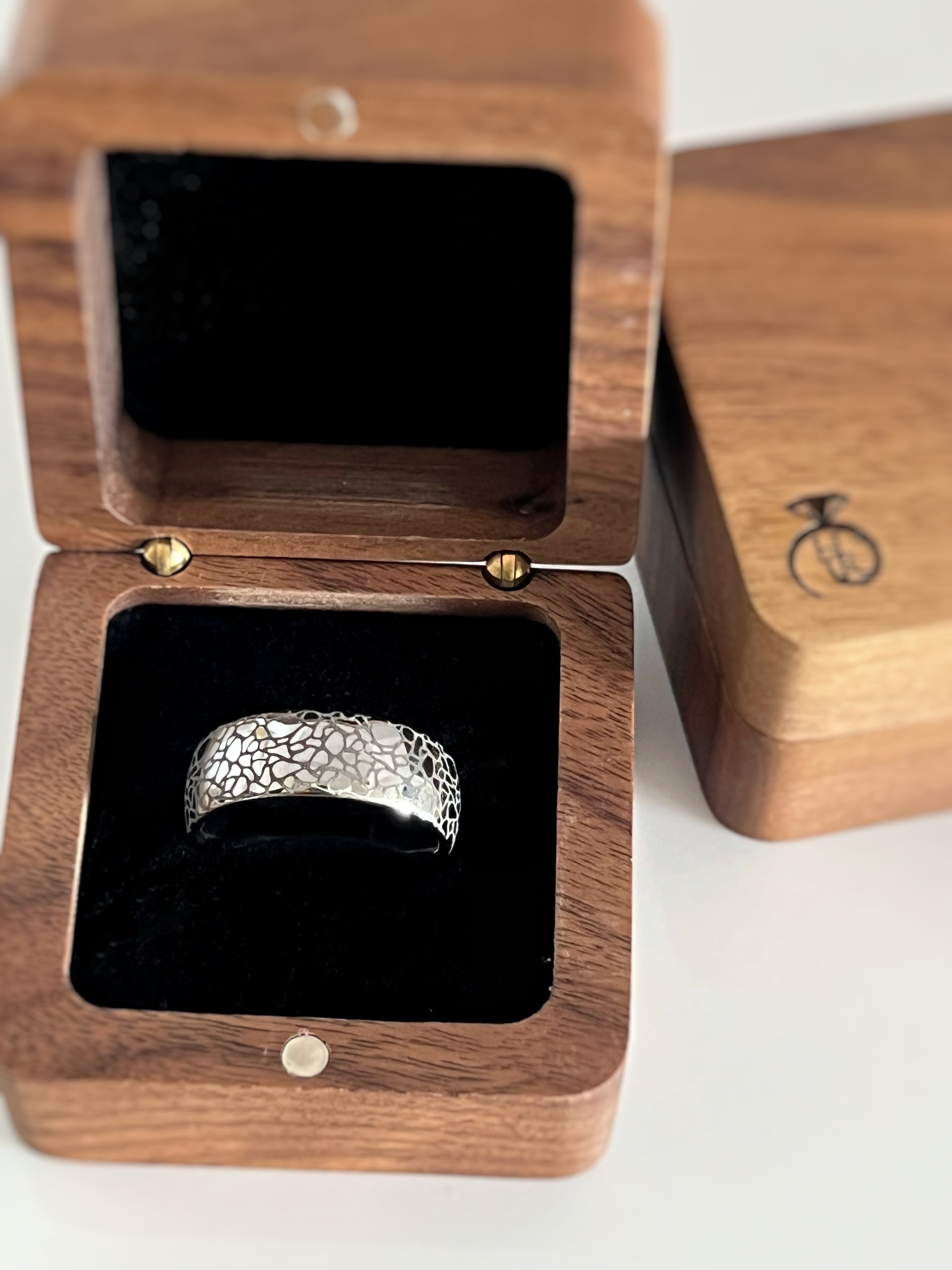 White gold tungsten ring with a cracked earth pattern, displayed in an open wooden ring box with a black interior.