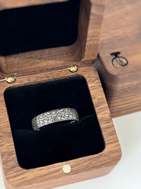 Silver tungsten ring with intricate black botanical leaf engraving, displayed in a wooden ring box. Elegant nature inspired wedding band with detailed floral pattern.
