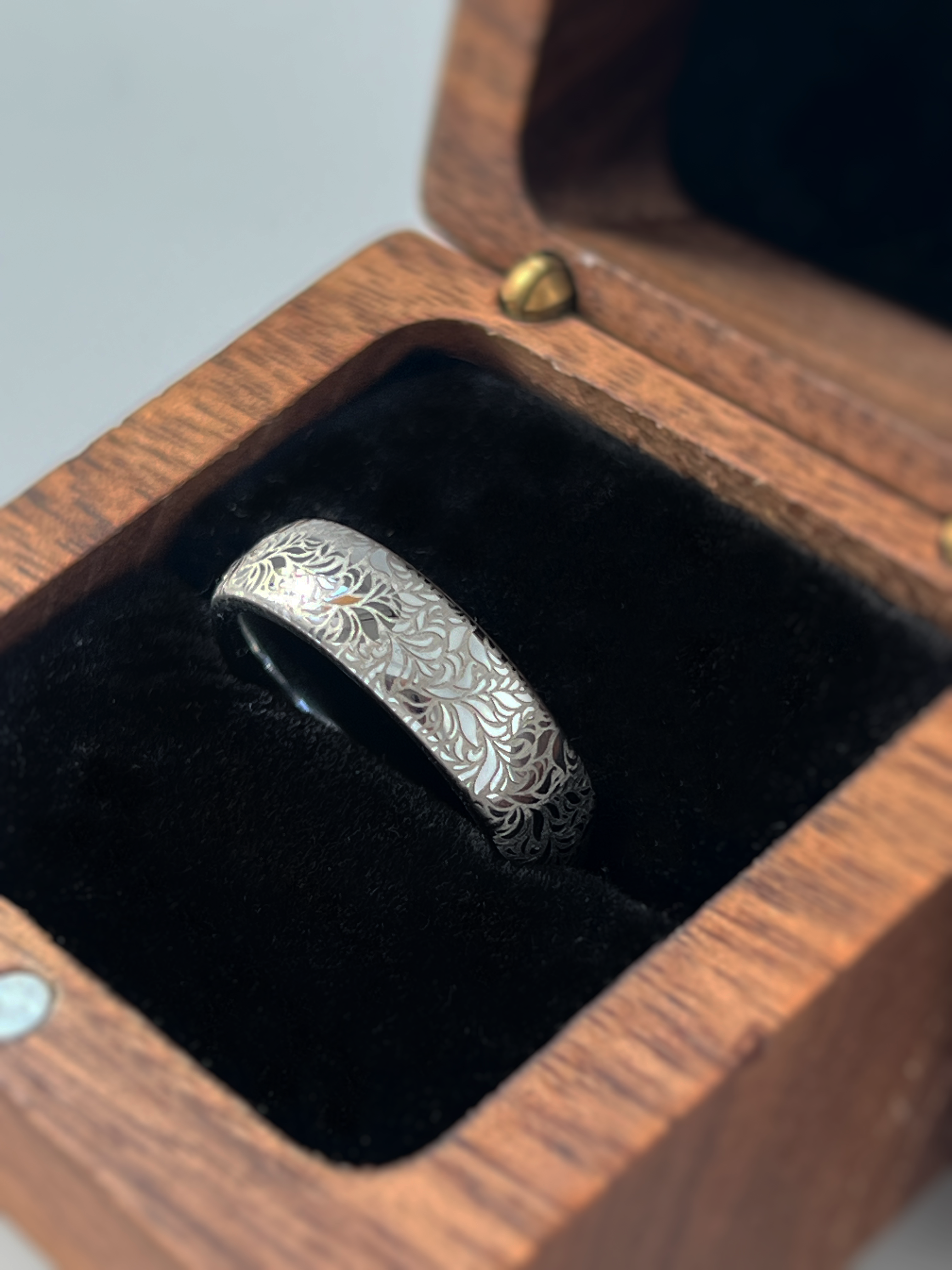 Elegant white gold tungsten displayed inside wooden box. The ring has  intricate silver floral engraving, featuring a polished shiny finish and delicate patterns. Durable and stylish, this engraved tungsten wedding or engagement band for her or him