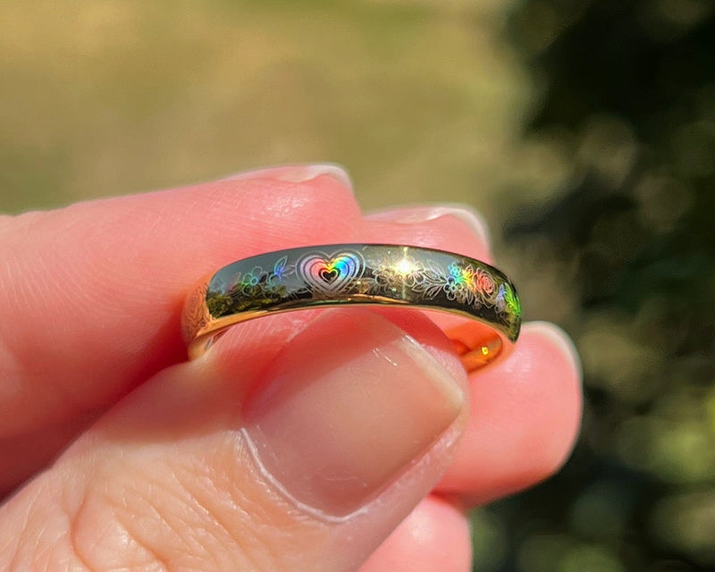 Heart in Bloom ring in a gold surface color, held between fingers in sunlight, with a holographic heart and floral pattern reflecting rainbow colors.