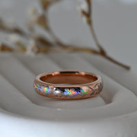 Rose-gold Hibiscus Glow tungsten ring with holographic hibiscus flowers and flowing leaves, resting on a pale display surface.