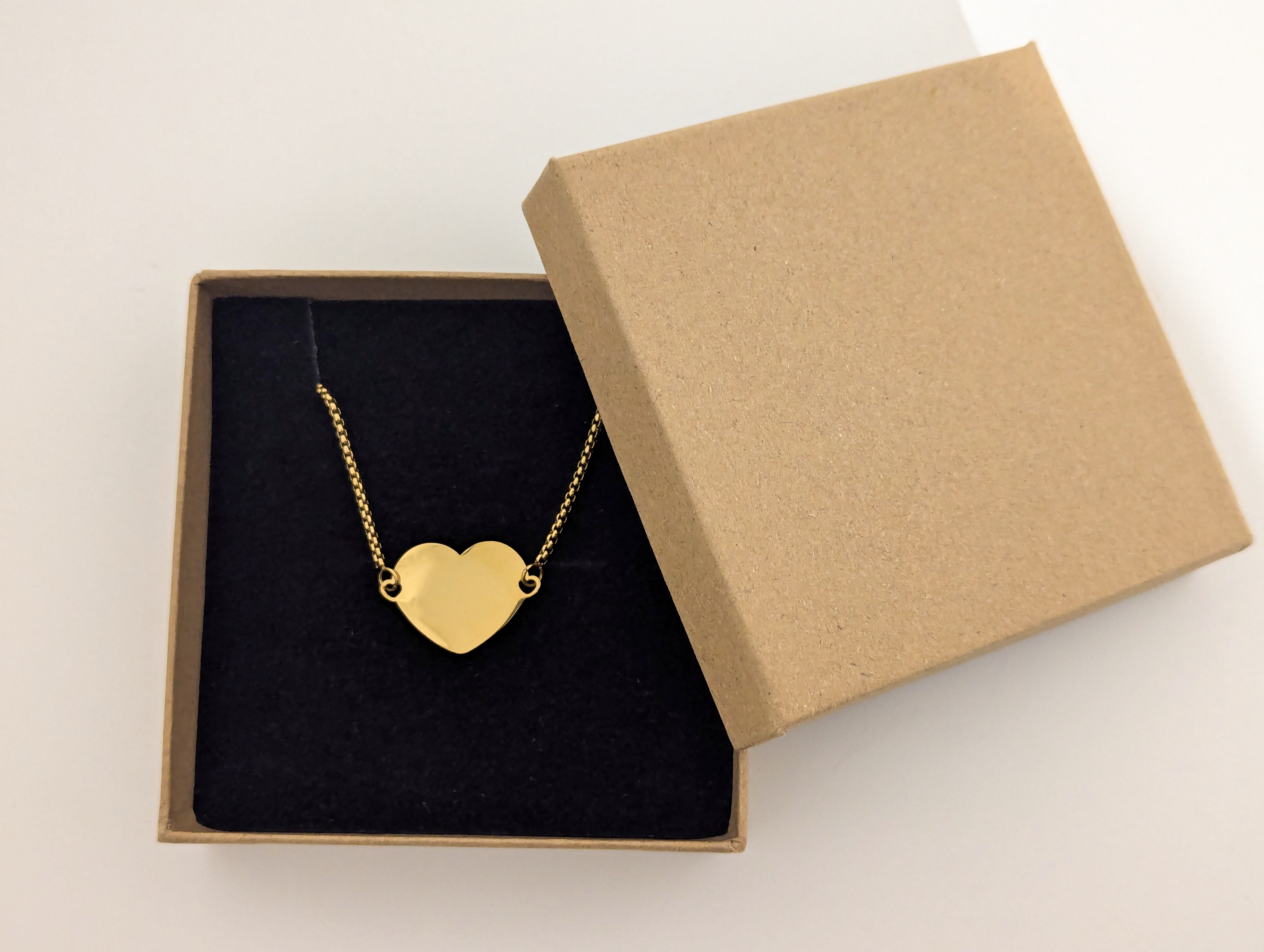 A gold-plated stainless steel heart-shaped bracelet displayed in a simple brown gift box with a black velvet lining. The bracelet features a smooth, unengraved surface, ideal for customization or as a timeless and elegant accessory. The durable gold-plated chain enhances the polished and sophisticated design.