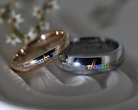 Two wedding bands displayed on a white sculptural stand, one rose gold and one silver, both featuring the word 'community' in rainbow-colored lettering.