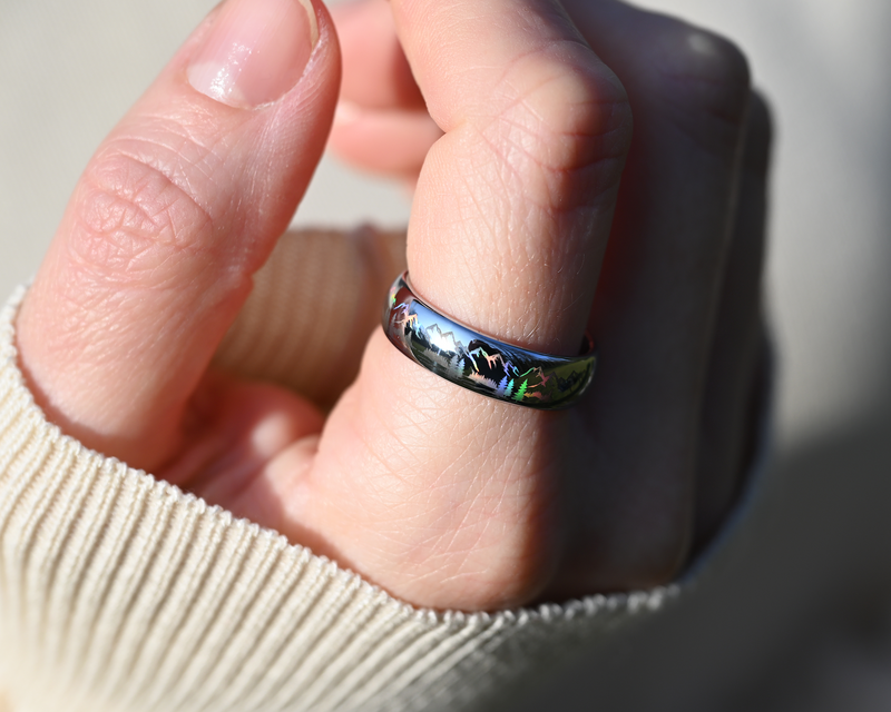 Close-up of the Radiant Mountains holographic tungsten ring worn on a hand, showing mountain, forest and wildlife silhouettes with rainbow reflections.
