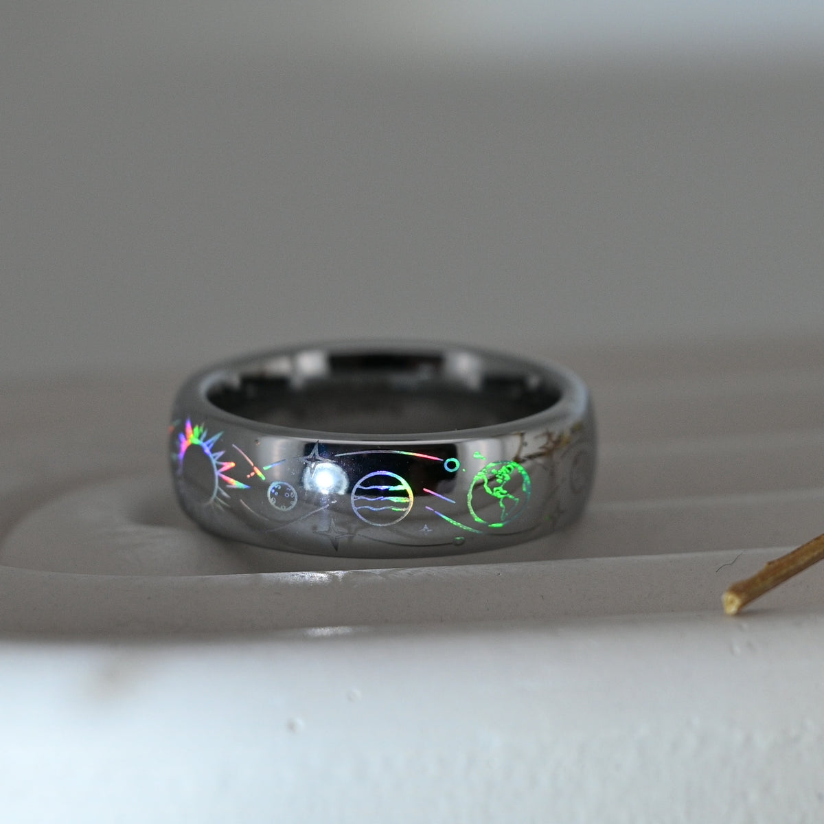 Silver-tone Celestial Orbit tungsten ring with holographic planets, stars and orbital lines, resting on a pale display surface.