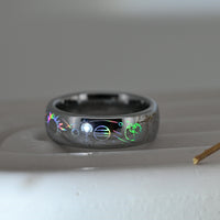 Silver-tone Celestial Orbit tungsten ring with holographic planets, stars and orbital lines, resting on a pale display surface.