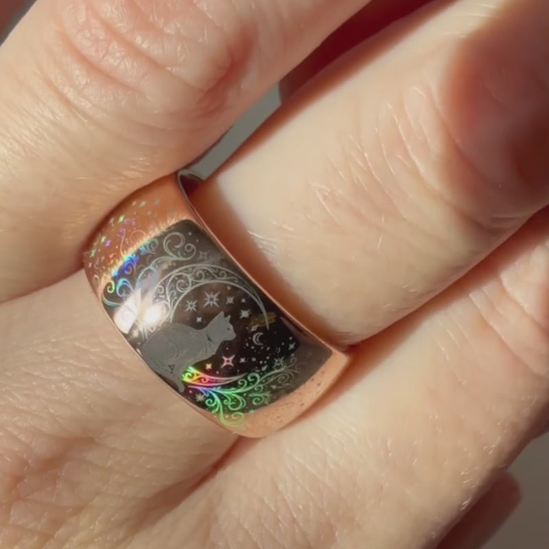 Holographic tungsten ring worn on hand, featuring a black cat surrounded by stars and celestial patterns with shifting rainbow reflections on a polished rose gold band.