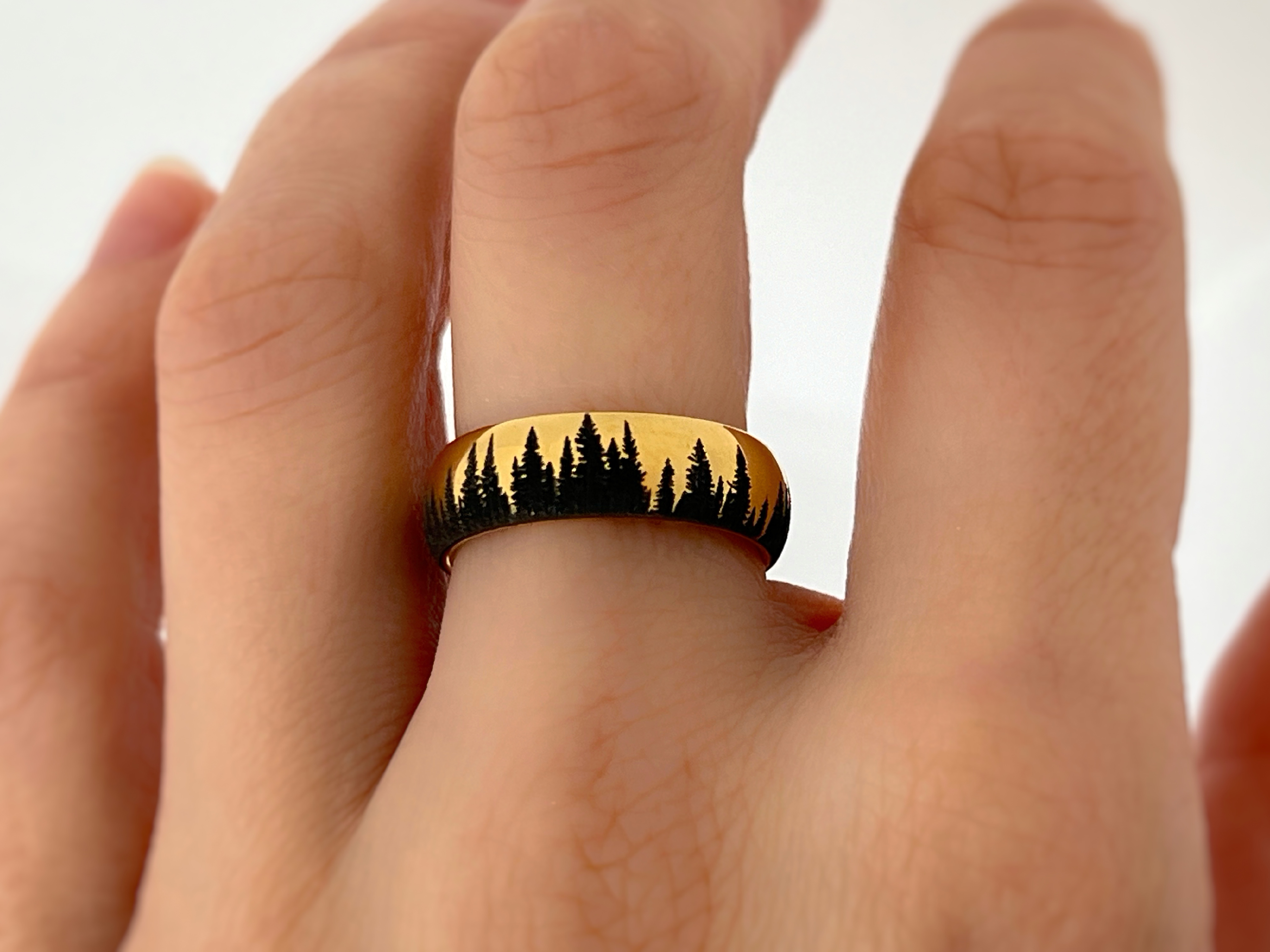 Gold tungsten ring with an engraved silhouette of an evergreen forest, worn on a finger, showcasing the detailed forest design.